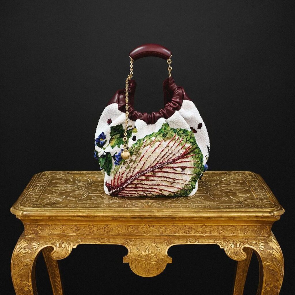 Loewe Squeeze Bag sa listom salate na beloj površini i braon drškom, sa crnim zidom iza nje i na zlatnom stolu